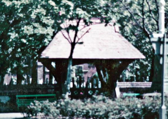 A lychgate showing a church graveyard and many trees in the background on either side of the gate are benches.