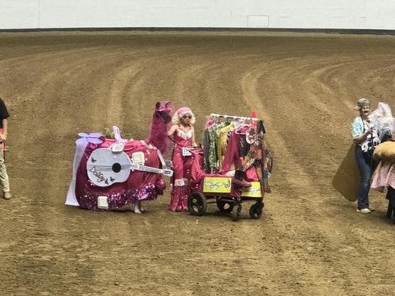A llama dressed up as Dolly Parton with a guitar and everything. The handler is also dressed like Dolly and carting a costume cart 