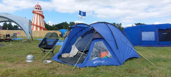 A blue dome tent with a camping chair and trangia stove next to it. In the background an orange and white helter skelter against a blue sky with some light white cloud. 