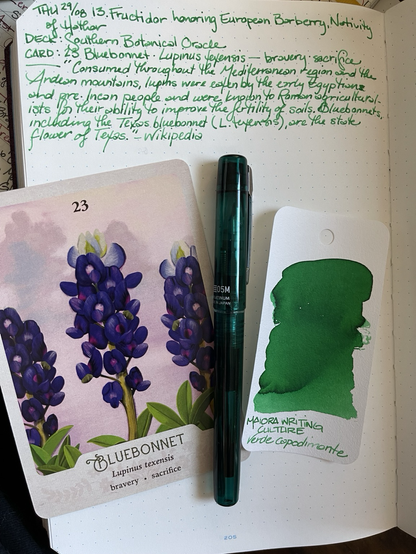 Open notebook with handwritten text from the post at the top of the page with additional text: —“Consumed throughout the Mediterranean region and the Andean mountains, lupins were eaten by the early Egyptian and pre-Incan people and were known to Roman agriculturalists for their ability to improve the fertility of soils. Bluebonnets, including the Texas bluebonnet (L. texensis), are the state flowers of Texas. — Wikipedia” below the text is a large format card featuring a digital painting of the upright flower stalks of three bluebonnets with a smattering of leaves across the bottom of the image. Next to the card is a clear dark green plastic fountain pen with a silver tone clip. Next to the pen is an ink swatch card showing a standard emerald green ink. 