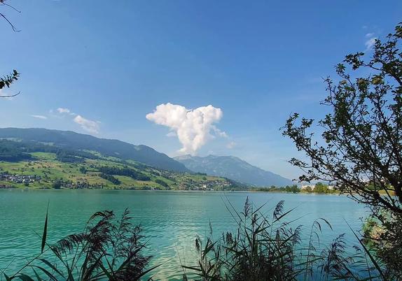 Blick über den See, der Himmel ist blau, wie auch das Wasser. Im Hintergrund sind Berge zu sehen. In der Bildmitte über einem Berggipfel ist eine Wolke, aus deren Form ein Hund herauszulesen ist. 