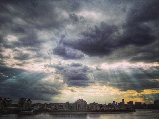 Dramatisch dunkle Wolken lassen ein paar Sonnenstrahlen durch und beleuchten Häuser der Isle of Dogs in London, im Vordergrund die Themse. 
