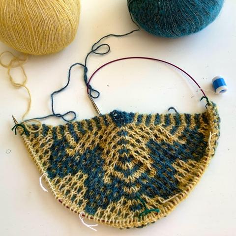 A triangular shawl with mosaic patterning being knit on a circular needle. It’s shown with a ball of yellow and a ball of petrol colored yarn on a white background.
