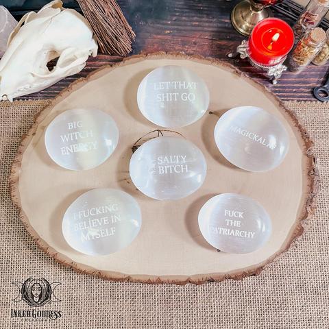 Six versions of the "Snarky Selenite Palm Stone" product are arranged on a raw wood slice. The phrases engraved on each of them include "LET THAT SHIT GO," "BIG WITCH ENERGY," "MAGICKAL AF," "SALTY BITCH," "I FUCKING BELIEVE IN MYSELF," and "FUCK THE PATRIARCHY." Presented by Inked Goddess Creations.