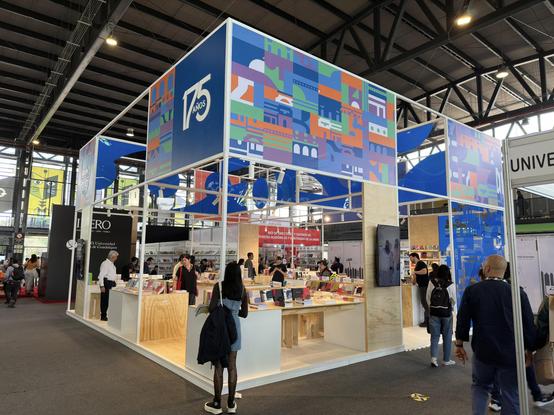Udelar's stand at the book fair. It's colorful, with wooden floors, tables, and benches. Books are displayed on the tables. Above, the Udelar logo and "175 years" are visible.