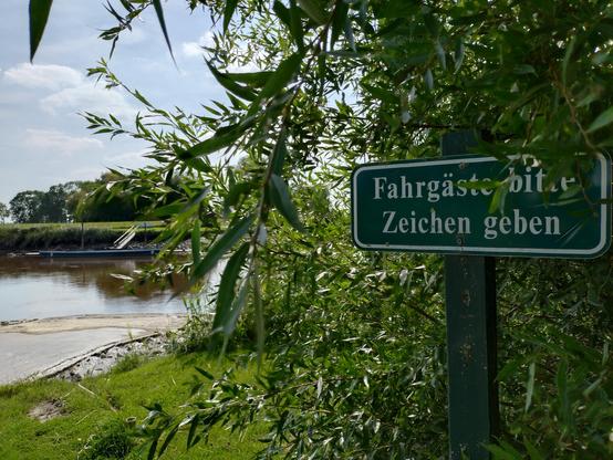 Auf dem Bild sieht man.ganz nah auf der rechten Seite ein grünes Schild mit weißem Rand und weißer Schrift: Fahrgäste bitte Zeichen geben. Drum herum sind Büsche.
Im linken Teil des Bildes sieht man eine gepflasterte Rampe hinunter zu einem Wasserlauf. Auf der gegenüberliegenden Seite liegt eine blau gestrichene, flache Fähre in Hand-Betrieb vor einem kleinen Deich.