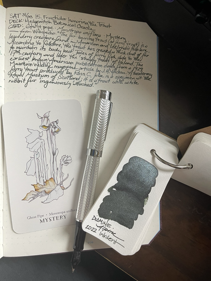 Open notebook with handwritten text from the post at the top of the page with additional text: —from Wikipedia: “The fur-bearing trout (or furry trout) is a legendary creature found in American folklore and Icelandic folklore. According to folklore, the trout has created a thick coat of fur to maintain its body heat. Tales of furry fish date to the 17th-century and later the "shaggy trout" of Iceland. The earliest known American publication dates from a 1929 Montana Wildlife magazine article by J.H. Hicken. A taxidermy furry trout produced by Ross C. Jobe is a specimen at the Royal Museum of Scotland; it is a trout with white rabbit fur "ingeniously" attached.” Below the text is a card with a pen & ink drawing of ghost pipe plants with very minimal coloring. Next to the card is an uncapped silver-tone fountain pen with wave pattern. Next to the pen is a swatch booklet on a ring opened to a dark olive ink with very heavy silver shimmer. 