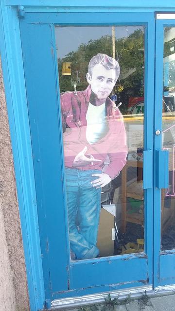 Colour photo looking at a blue set of doors with inset glass. The door on the left has a cutout of Jim Stark, the character played by James Dean in "Rebel Without A Cause". The pose we see captured in the cutout is one where Jim has his left hand over his left blue jeans front pocket, and his right hand is held above (in front of Jim's mostly unbuttoned red shirt with white undershirt beneath), with the index finger pointing to the right, and the thumb held nearly vertical.
