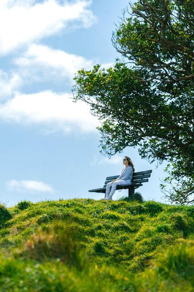 Woman sitting on a bench on top of a hill wearing sunglasses on a clear sky day, in a contemplative manner.