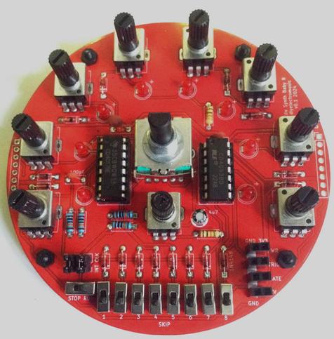 Photo of a red, circular PCB with 8 pots and LEDs around the edge, a pot and rotary switch in the middle, and a set of slider switches at the bottom.  The PCB is fully populated and there are two ICs near the middle.