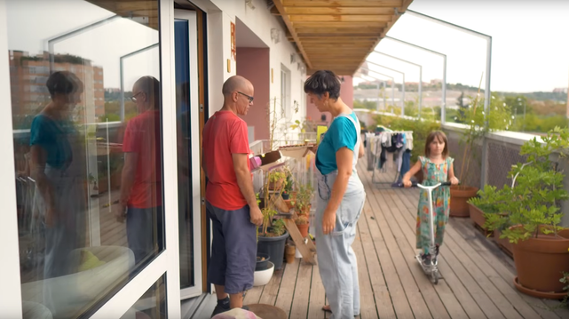 Fotograma del documental. Se ve un pasillo exterior común, con suelo de tarima, ropa tendida y plantas, con dos adultos hablando delante de la puerta de una casa, y una niña pasando en patinete.