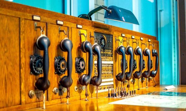 The photo shows a vintage telephone switchboard made of wood, featuring multiple rotary dial telephones with black handsets hanging on hooks. The switchboard includes various knobs, dials, and plugs used by an operator to manually connect calls. A lamp above illuminates the setup, likely in a historical or museum display. The image, with warm tones highlighting the wood, evokes a sense of nostalgia for early telecommunications technology. @HaroldSinnott