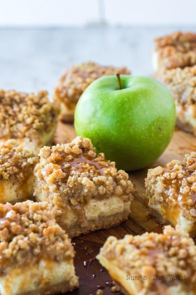 Sliced caramel apple cheesecake bars laid out on a cutting board with a single granny smith apple in the middle. Each caramel bar is topped with a crumble topping and caramel drizzle.