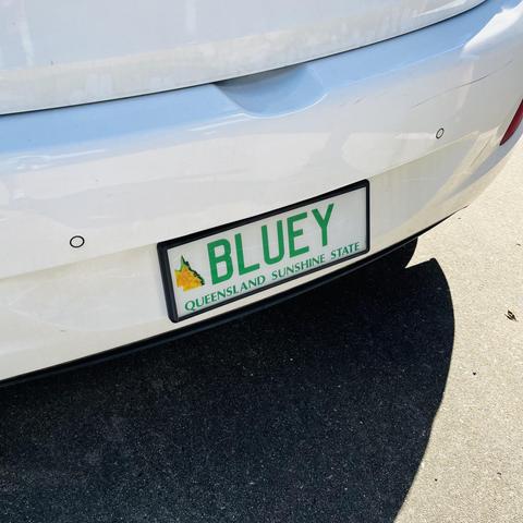 The front bumper of a white car showing a green-on-white “Queensland Sunshine State” personalised number plate featuring a topographical style map of Queensland on the left and an identifier of “BLUEY”