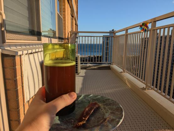 Closeup of a green glass filled with brown beer and a plate with a slice of dried fish in the foreground. The background is a highrise balcony with the Atlantic Ocean peeking in the background through the balcony railing
