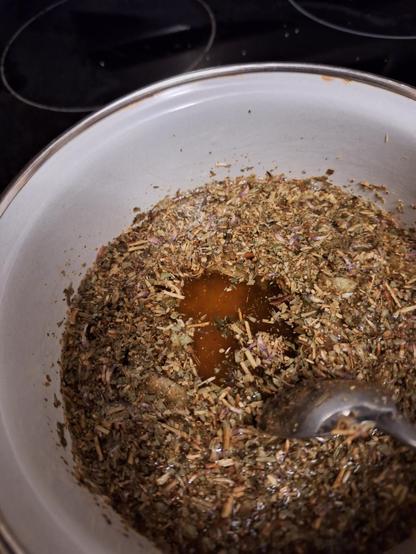 Herbs float on water in a white pot. A spoon makes a space to see the golden brown color the water has become