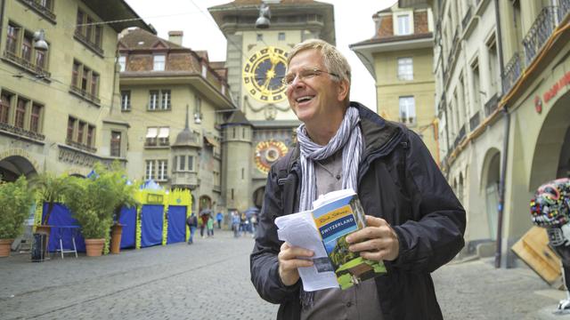 Rick Steves, white man with glasses and salt-and-pepper hair, wearing a scarf and a dark jacket, holding a blue and yellow guidebook. He is standing in a European town square with a big building with a gold clock in the background.