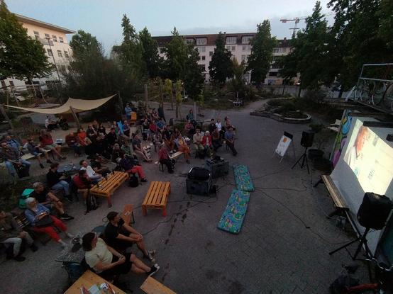 Foto von oben im Abendlicht auf einen Platz mit vielen Menschen, die vor einer Leinwand an einem Container sitzen (rechts im Bild). Im Hintergrund stehen Bäume und um den Platz Häuser. Die Menschen sitzen auf Bänken aus Paletten, Bierbänken, Tischen und Mauern vor einem Beet.