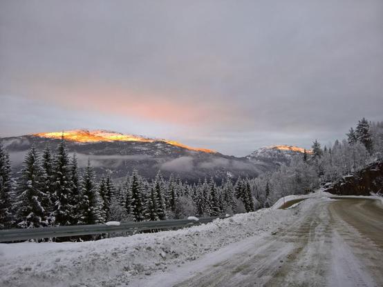 Ich stehe am Straßenrand. Die Straße ist vereist, hinter der Leitplanke sind schneebedeckte Nadelbäume zu sehen. In der Ferne ist eine schneebedeckte Bergspitze, die in Geld- und Orange-Tönen leuchtet, weil sie von der untergehenden Sonne angestrahlt wird.