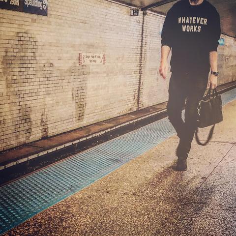 A person walking down a subway platform wearing a black tee shirt with the words “whatever works” in white block letters. They are carrying a satchel of some sort — it appears to be a computer case. 