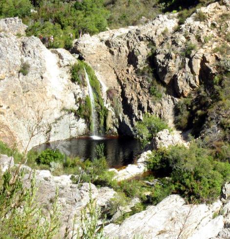 A photo of a waterfall falling into a round pool from above.
Rocks and sparse vegetation surround the pool. The water is a reddish brown caused by the fynbos vegetation the water moves through
A path with people on it is visibke above the pool and crossing the top of the waterfall
