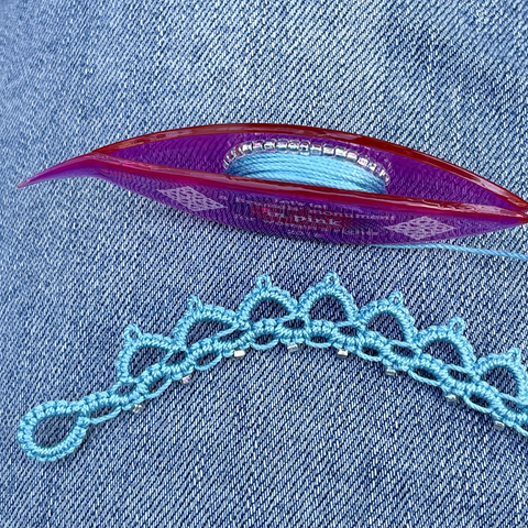 The start of a piece of tatting in light turquoise blue thread displayed on a blue jeans pant leg, with a reddish pink tatting shuttle wrapped with thread and silver lined seed beads.