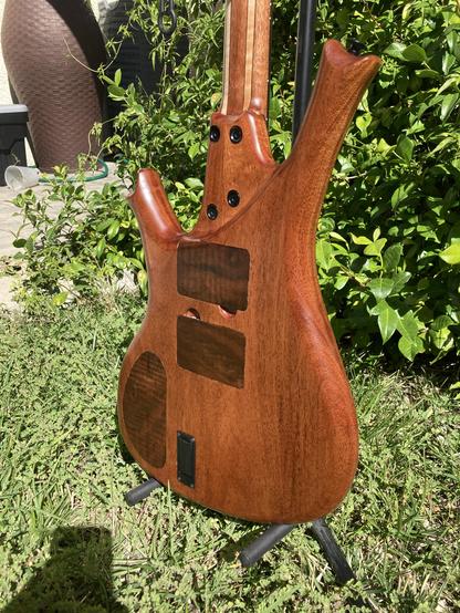 A sunlit baritone guitar sits with its back showing, on a stand in a yard, with foliage behind it.