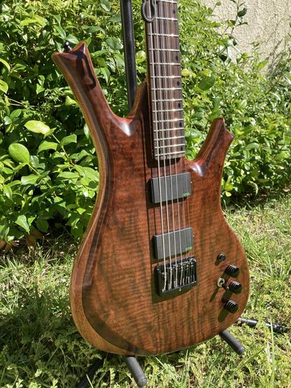 A sunlit baritone guitar sits on a stand in a yard, with foliage behind it.