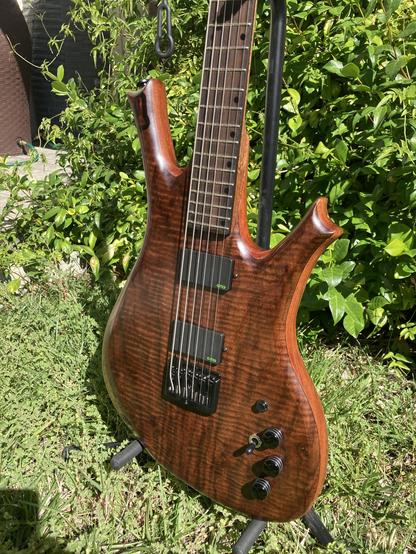 A sunlit baritone guitar sits on a stand in a yard, with foliage behind it.