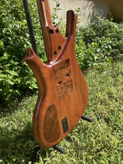 A sunlit baritone guitar sits with its back showing, on a stand in a yard, with foliage behind it.