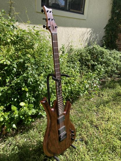 A sunlit baritone guitar sits on a stand in a yard, with foliage behind it.