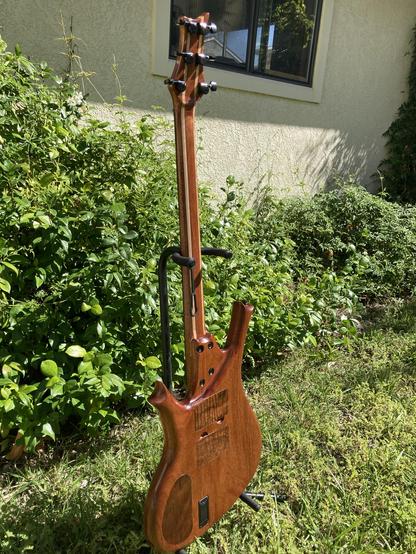 A sunlit baritone guitar sits with its back showing, on a stand in a yard, with foliage behind it.