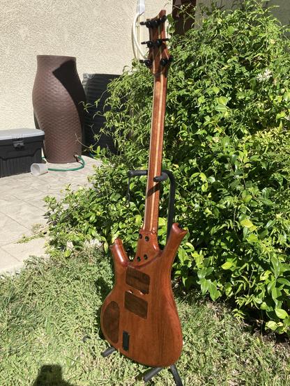 A sunlit baritone guitar sits with its back showing, on a stand in a yard, with foliage behind it.