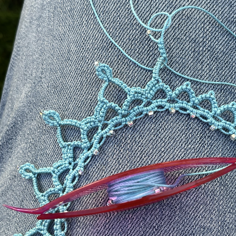 Tatted lace in progress in light blue thread, forming a design with points. Tatting shuttle in pinkish red color is carrying some of the thread.