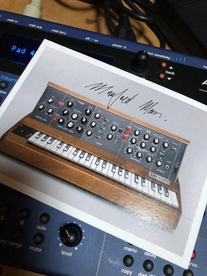 Unterschrift von Manfred Mann auf einer Postkarte mit Minimoog-Motiv, dem Synthesizer, den er jahrzehntelang virtuos nutzte. Inzwischen hat er ihn gegen verlässlicheres Equipment eingetauscht. Für Soli nimmt er den Novation Peak, angesteuert über eine von zwei Novation X-Stations.