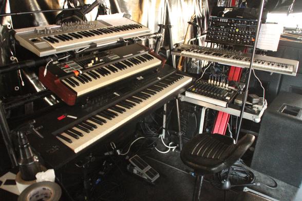 Die Keyboardburg von Manfred Mann. Im rechten Winkel zueinander stehen ein dreifach Rig mit Novation X-Station, Hammond XK-1c und Rolad RD-800, sowie rechts davon ein Rig mit Novation Peak, einer weiteren Novation X-Station sowie einer Ablage für Submischer und Gedöns.