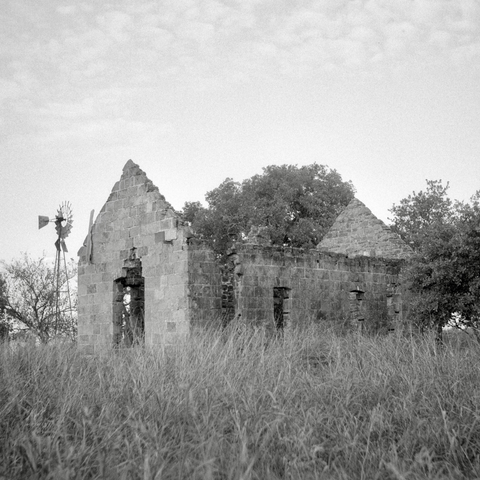 A black and white film photo. An abandoned stone house sits in a field with an old windmill in the background in Pontotoc, Texas. Sunday, September 1, 2024.