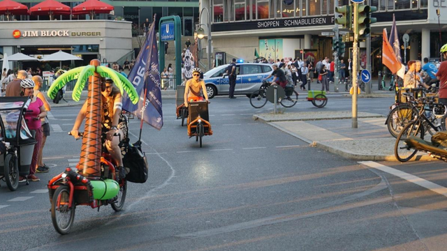 Diverse Lastenradfahrende und adfahrende mit Anhänger umkreisen die Mittelinsel auf der Kantstrasse gegenüber des Bikini Berlin auf der September Demo.
Im Vordergrund ein Lastenrad, beladen mit einer aufgeblasenen Palme.
Im Hintergrund ist ein weiteres Lastenrad, sowie ein Fahrrad mit Anhänger zu sehen.