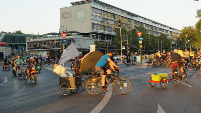 Diverse Lastenradfahrende und adfahrende mit Anhänger umkreisen die Mittelinsel auf der Kantstrasse gegenüber des Bikini Berlin auf der September Demo.
Im Vorfergrund sieht man leicht verschwommen ein Fahrrad mit Anhänger, worin ein großer weißer Teddy mit Sonnenhut sitzt, Rechts ein Fahrrad mit kleinme Transportanhänger und links im Hintergrund fahren weitere Fahrräder verschiedener Bauart auf dei Kamera zu.