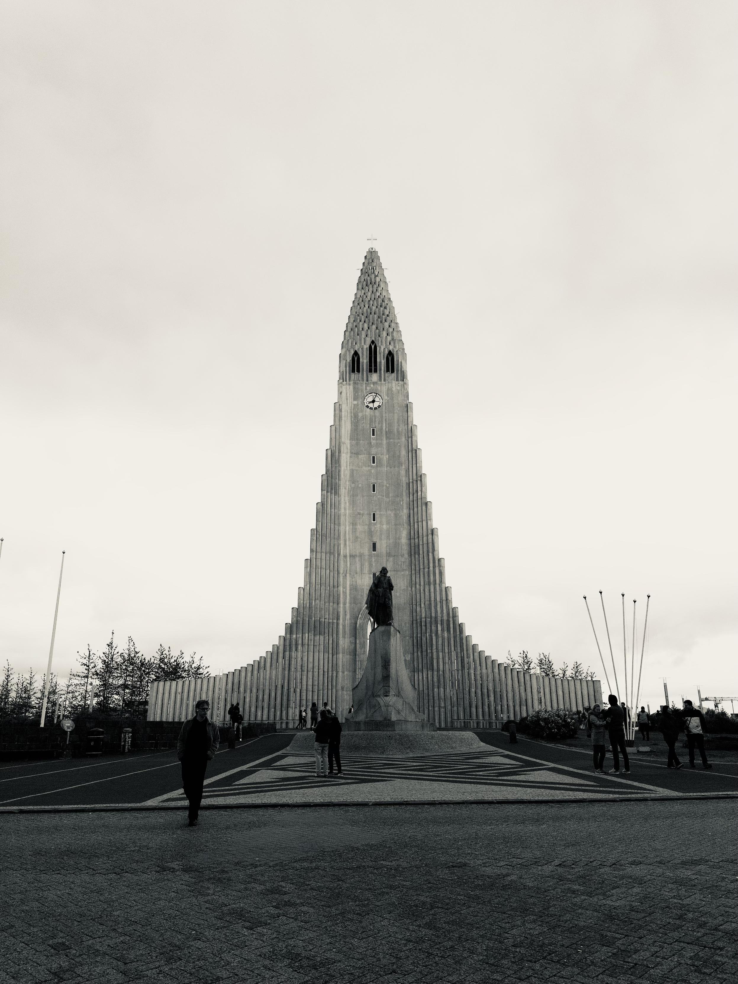 Hallgrímskirkja on a gloomy day in black and white. Some tourists can be seen out front as well as a statue of Lief Eriksson. 