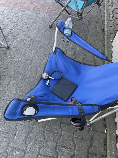 Campingstuhl in der Boxengasse im Schatten mit Wasserflasche, Luftdruckmessgerät, Ohrschützern und ebook Reader.