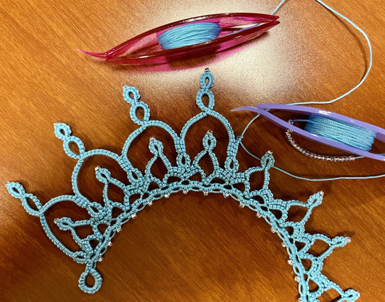 Tatted lace in progress showing a scalloped collar design with points. 2 tatting shuttles are attached with the light blue thread used in the project. 