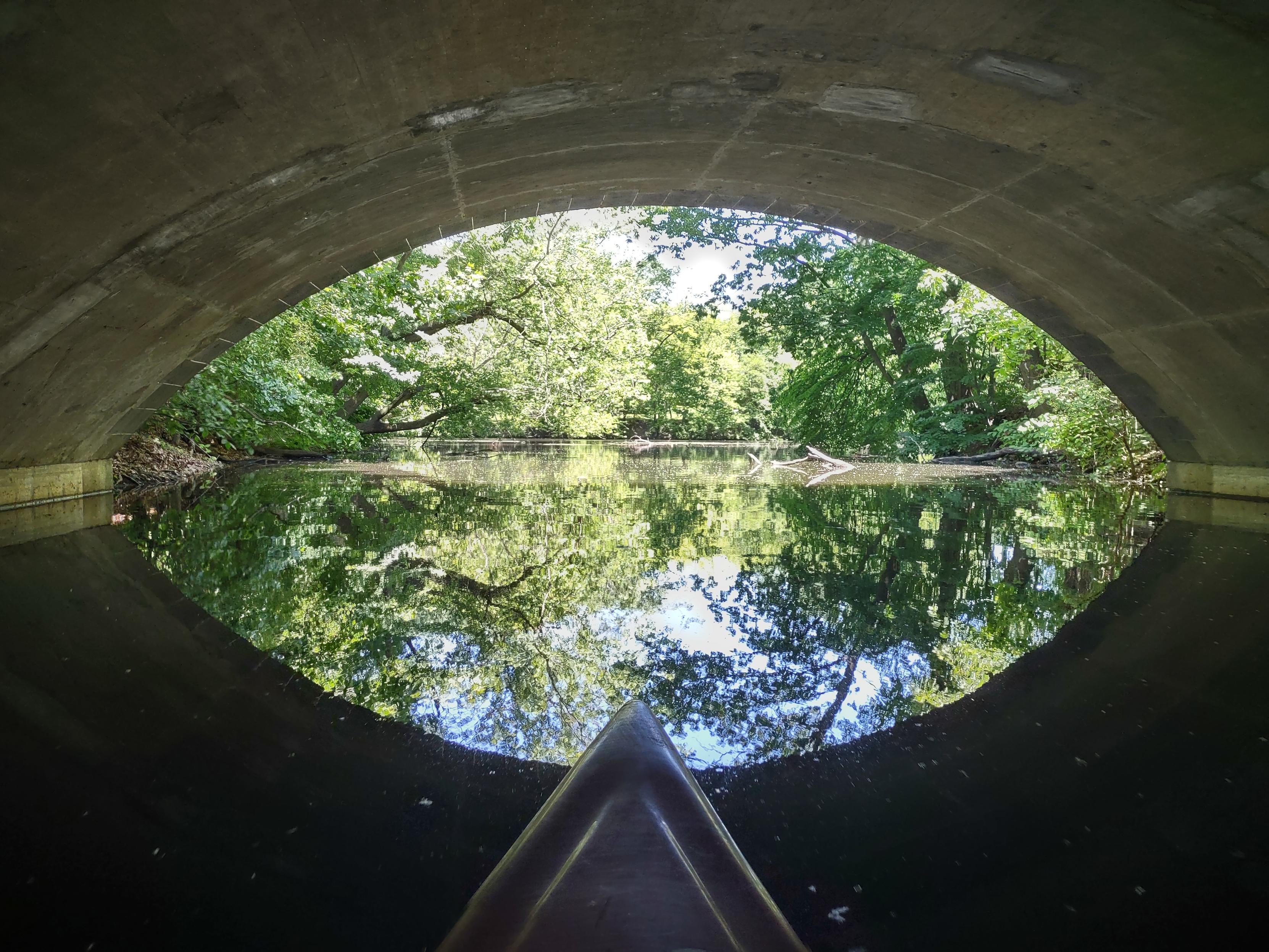 The view under a curved bridge, with a tree-lined stream reflected: an bright eye-shaped study in blue and green, surrounded by the dark underside of the bridge. The tip of my kayak is at the bottom.