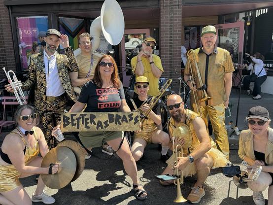A lively group of musicians poses outdoors, wearing gold outfits and sunglasses. They hold various instruments, including trumpets and a tuba. A woman in the center holds a sign reading "BETTER AS BRASS." The setting appears festive and colorful,