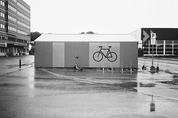 Svart hvit foto av et sykkelskurbygning. Bygningen har et stort sykkelsymbol. Noen få elsparkesykler er parker rundt. Asfalten er våt av regn.