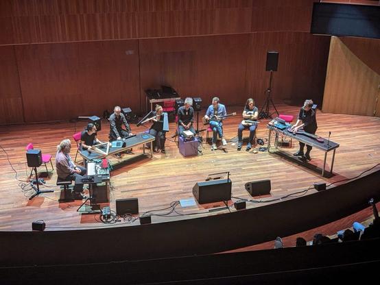 A group of eight musicians is performing on stage in a concert hall, featuring various instruments and electronic equipment. The setting includes wooden walls and a semi-circular seating arrangement, with some audience members visible. The musicians appear focused on their performance.