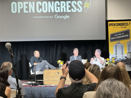 From left-to-right: Andy Langer, Lyle Lovett, Terry Lickona. On stage at 800 Congress for the Texas Tribune festival. In the foreground, audience members and a microphone.