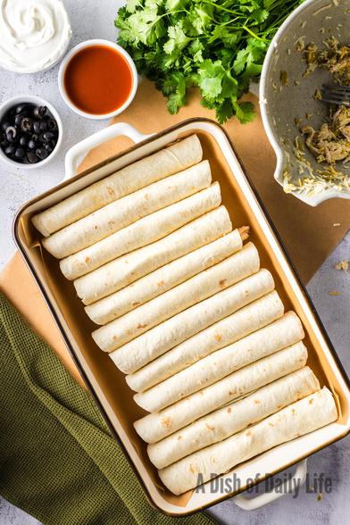 12 Wrapped and rolled enchiladas in a pan.  Nothing is on top of the enchiladas (Yet!) with various toppings all around in photo and a green dish towel. 