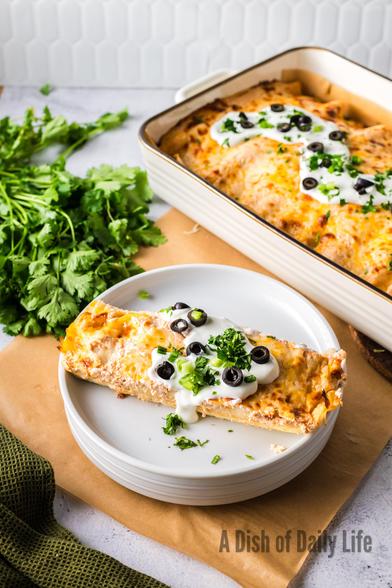 Baked Enchiladas in casserole dish in the background. In front are two enchiladas topped with sour cream, black olives, green onions and cilantro on a plate. 