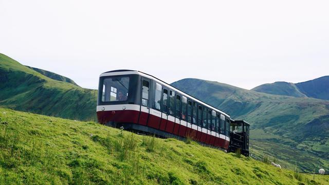 Bergbahn (Snowdon Mountain Railway) im Vordergrund mit grünem Gras. Im Hintergrund grüne Berge und diesiger Himmel.